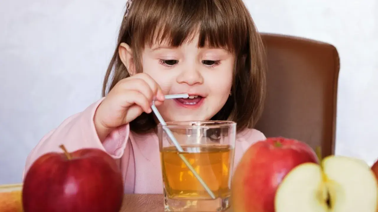 Mała dziewczynka pije sok jabłkowy, który pomaga na zaparcia. Obok stoją jabłka.