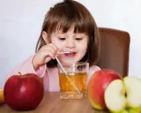 Mała dziewczynka pije sok jabłkowy, który pomaga na zaparcia. Obok stoją jabłka.