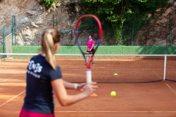 Dwie kobiety grają w tenis ziemny na korcie. Jedna z nich, z rakietą w ręku, przygotowuje się do uderzenia.