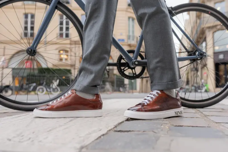 Eleganckie sneakersy do garnituru w brązowym kolorze, zestawione z szarymi spodniami, na tle miejskiego krajobrazu z rowerem.